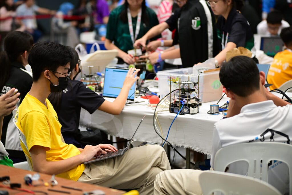 Hwa Chong Institution student working on a RoboCup Junior Soccer robot at RoboCup Singapore Open 2023, with Raffles Girls’ School students in the background.
