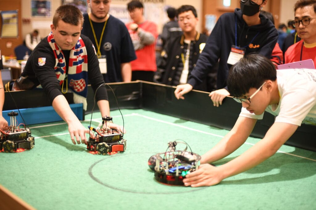 Teams preparing autonomous soccer robots on a green field at RoboCup Asia-Pacific 2023 in PyeongChang.